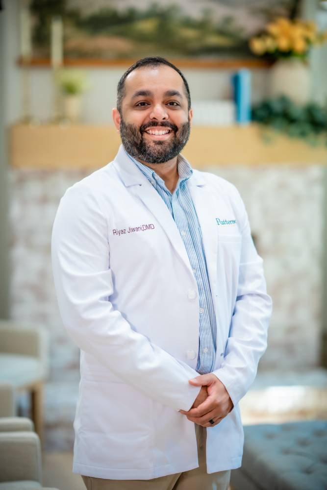 Dr. Riyaz Jiwani, DMD, smiling in a white lab coat over a striped collared shirt at Patterson Family Smiles in Clayton, GA 