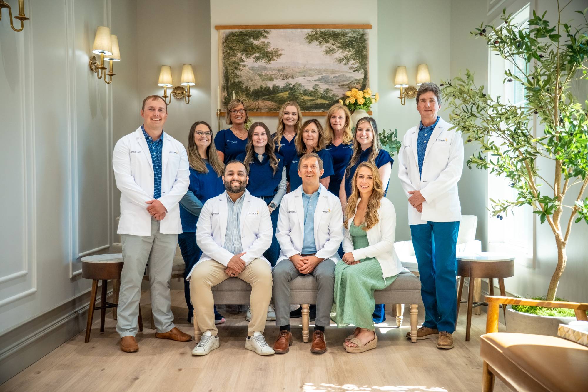 Patterson Family Smiles dental team of five dentists in white coats and staff in navy blue scrubs smiling together in the Clayton, GA office lobby with landscape artwork, brass sconces, and potted olive trees 